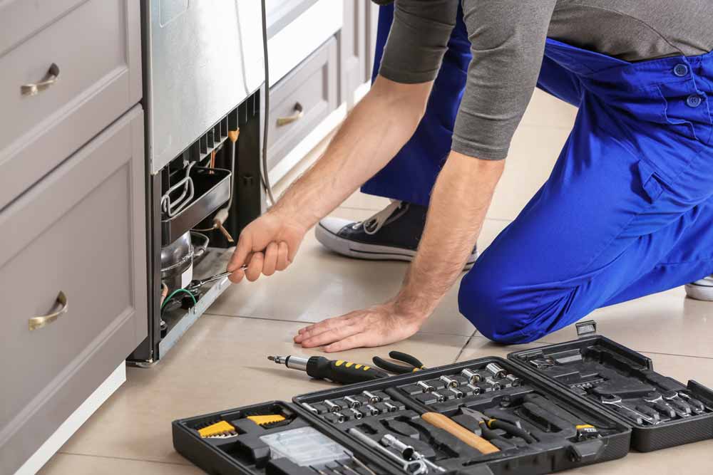 Repair man in blue overalls kneeling in front of refrigerator to repair it